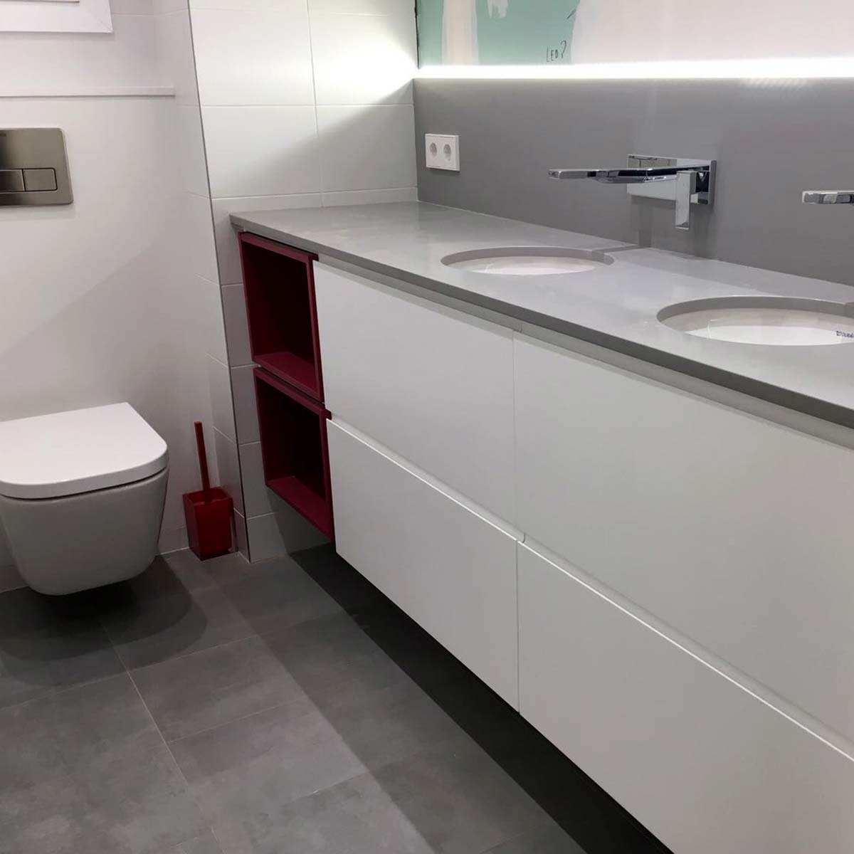 Bany amb WC i moble amb unes piques integrades de color blanc, amb un toc de vermell en les prestatgeries.